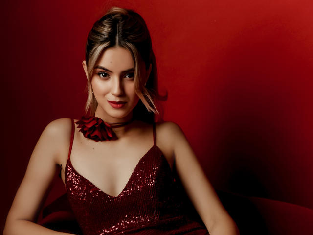 JasmineDay vestindo um vestido vermelho com lantejoulas e alças que envolvem o pescoço, sentada em frente a um fundo vermelho, sorrindo com batom vermelho, cabelo loiro em um rabo de cavalo, mostrando os seios..