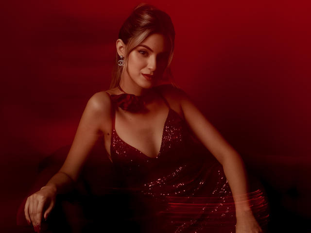 JasmineDay trägt ein rotes, paillettenbesetztes Kleid und zeigt dabei verspielt ihre Brüste, mit einem roten Hintergrund, Ohrringen und einer Halskette. Sie wirkt sexy und selbstbewusst..