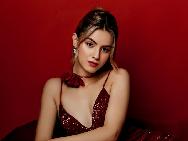 JasmineDay in einem roten, paillettenbesetzten Kleid, mit tiefem Ausschnitt, kleinen Brüsten, einem roten Choker mit Blume, rotem Lippenstift und Ohrringen, vor rotem Hintergrund, blickt mit leichtem Lächeln in die Kamera..