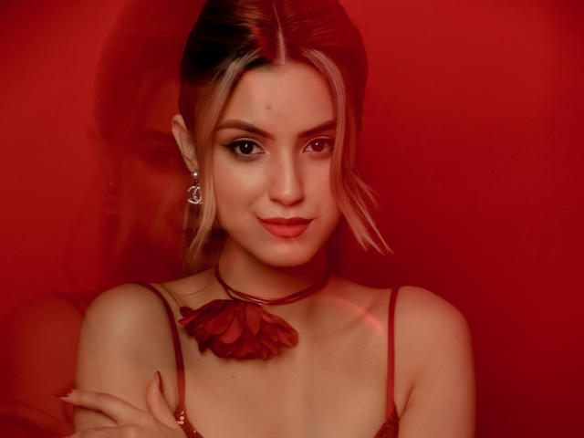 JasmineDay with red background, wearing red choker, flower necklace, and earrings, has light blonde hair, subtle makeup, and a cute smile, holding her shoulder with red-lit shadow.