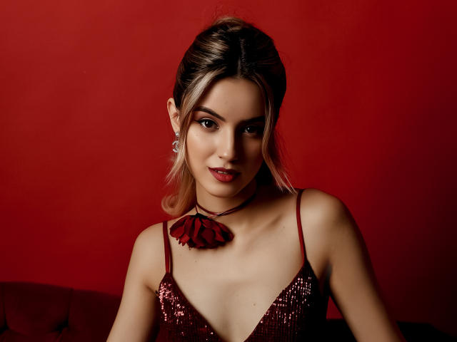 JasmineDay with blonde hair, wearing a red sequined dress, choker with flower, and earrings, smiling at the camera, red background, showing her tits and cleavage.