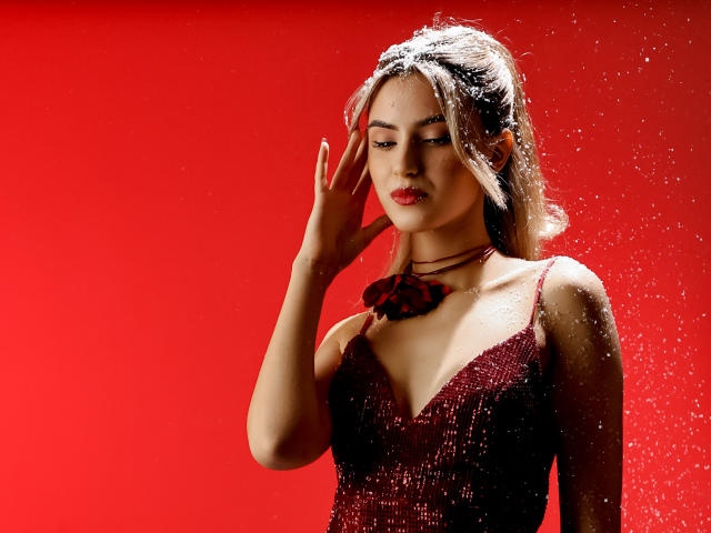 JasmineDay con cabello rubio claro, vestido rojo y lazo rojo, gotas de agua en su piel, mano cerca de la cara, mirando hacia abajo, fondo rojo, tiene un buen busto..