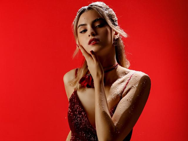 JasmineDay mit rotem Hintergrund, trägt ein glitzerndes rotes Kleid, Hand am Kinn, dezentes Make-up, rote Lippen, blondes Haar in Zöpfen, kleine Brüste, Choker, schimmernde Haut, selbstbewusster Ausdruck..