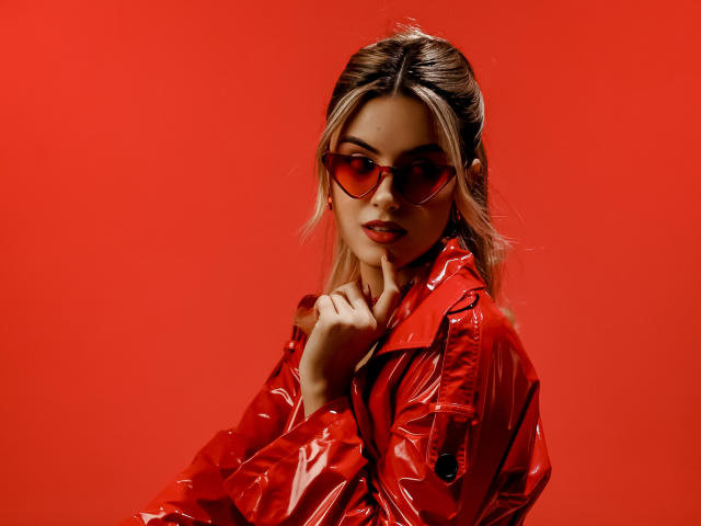 JasmineDay met een rode, glanzende jas, bijpassende rode zonnebril en blond haar, die haar kin aanraakt, met een verleidelijke blik, op een rode achtergrond. Borsten niet zichtbaar, focus op het gezicht en het bovenlichaam..