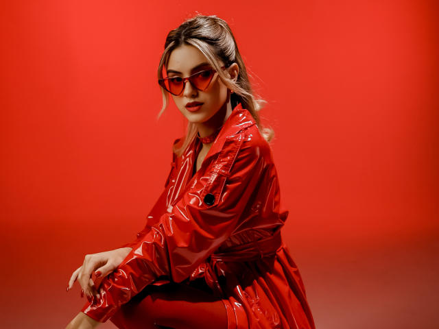 JasmineDay con gafas de sol de color rojo, vistiendo una chaqueta roja brillante de látex y cabello rubio recogido en una coleta, sentada con las manos en las rodillas, luciendo sexy y segura de sí misma..