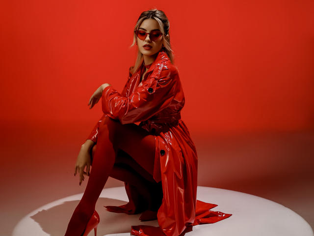 JasmineDay in een rode latex outfit, in een hurkpositie, met rode zonnebril, laarzen en hoge hakken, met glanzend, reflecterend materiaal, er verleidelijk uitzien, haar borsten en billen zijn licht zichtbaar..