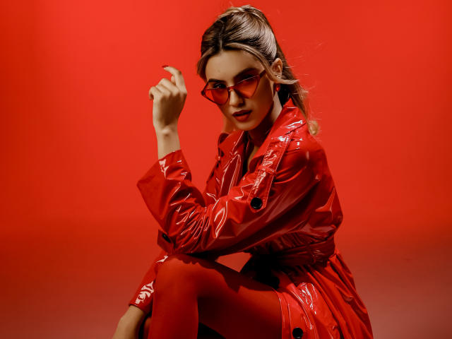 JasmineDay in shiny red vinyl coat, red tights, and red sunglasses, crouching with one hand near her face, looking sexy with her ass slightly raised.
