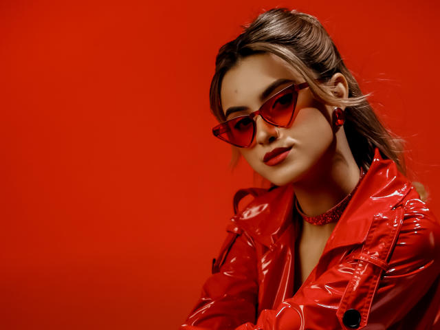 Jasmine, avec une veste rouge brillante, des lunettes de soleil rouges, du rouge à lèvres rouge et un collier rouge, a l'air élégante et confiante, et porte des boucles d'oreilles rouges, sur un fond rouge uni..
