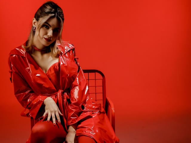 JasmineDay in einem glänzenden roten Lateikleid, sitzend auf einem Stuhl, roter Hintergrund, lächelnd, roter Choker, rote Nägel, mittelgroße Brüste, blondes Haar, blickt in die Kamera, Hände auf dem Stuhl..