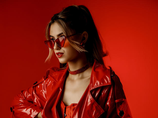 JasmineDay mit roter, glänzender Jacke, rotem Choker, roten, herzförmigen Sonnenbrillen und rotem Lippenstift, Blick zur Seite, dunkelbraunes Haar im Pferdeschwanz, roter Hintergrund, stilvoll und sexy..
