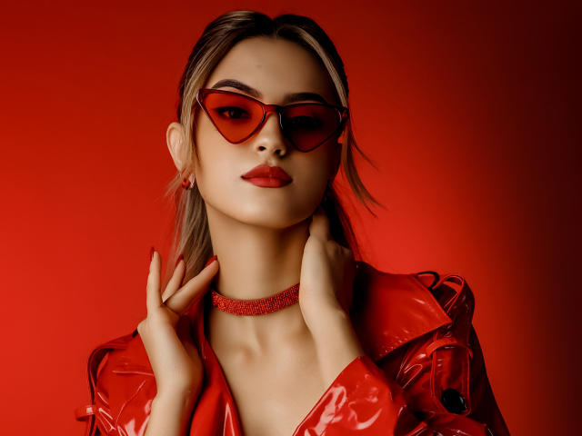 JasmineDay with red sunglasses, red lips, and red choker, wearing a shiny red leather jacket, touching her neck, hands up, blonde hair, red background, looking seductive.