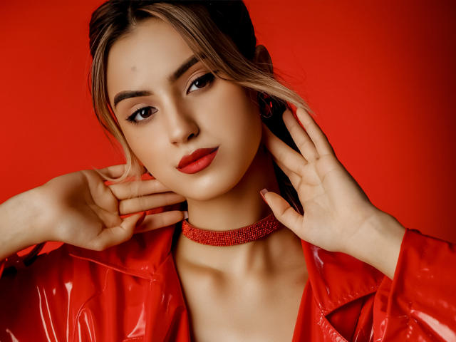 Jasmine mit rotem Lippenstift, trägt eine glänzende rote Jacke und eine rote Halskette, Hände am Hals, blondes Haar, roter Hintergrund, blickt in die Kamera, leicht lächelnd, hübsches Gesicht, kleiner Muttermal..