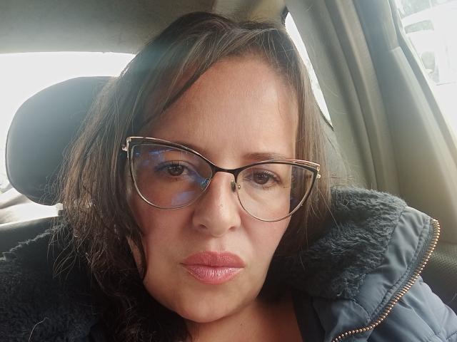 CandHoney hat braune Augen, mittelbraunes, welliges Haar und trägt eine Brille mit schwarzem Rahmen sowie eine schwarze, gepolsterte Jacke. Auf einem Foto ist sie in einem Auto zu sehen und man erkennt ihre rosafarbenen Lippen..