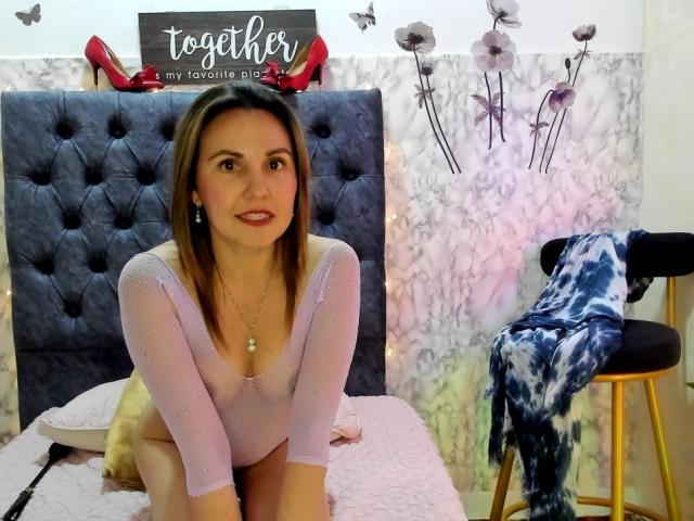 AdelaShery, with pink sparkly top, sitting on bed, showing ass, brown hair, red lipstick, small tits, necklace, bed with pink blanket, blue tie-dye scarf on stool.