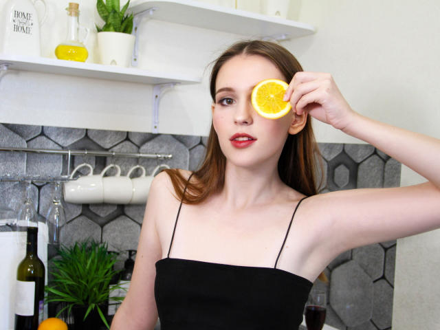 JuliaKing, avec une tranche d'orange sur son œil droit, portant un haut noir à bretelles fines, dans une cuisine avec des étagères blanches et un carrelage à motifs hexagonaux..
