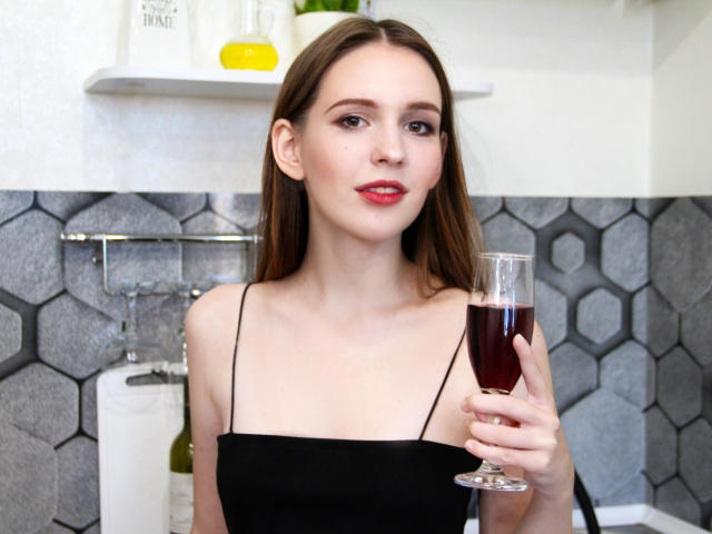 JuliaKing, com cabelos castanhos longos, batom vermelho, blusa preta sem alças, segurando um copo de vinho tinto, em pé em uma cozinha com azulejos hexagonais cinzas, olhando para a câmera..