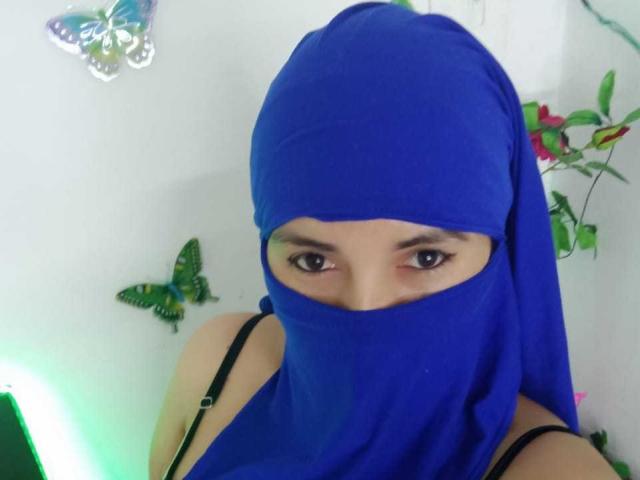 TrishaXx with blue headscarf covering her face and black bra straps visible, wearing a mask that exposes her eyes and eyebrows. Butterfly wall decorations in the background.