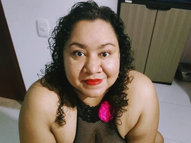 HiyunaMei, con i capelli ricci, rossetto rosso, top di rete nera e un fiore rosa, sorride alla fotocamera, mostrando le spalle scoperte e la parte superiore del petto, in una stanza leggermente buia..