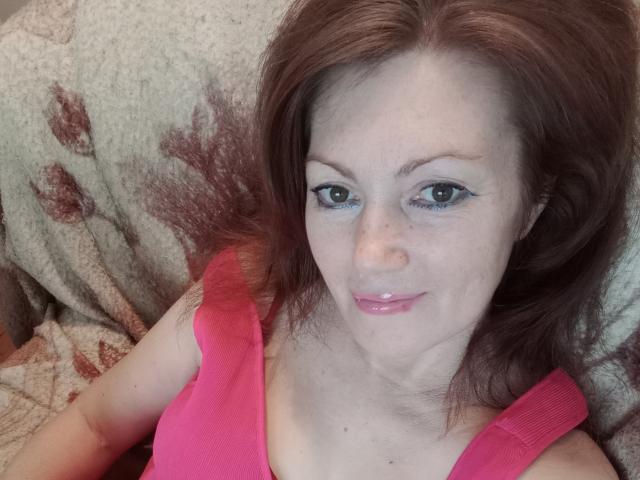SadieLorri, con cabello rojo, ojos azules y lápiz labial rosa, vestida con una camiseta rosa, sonriendo a la cámara, acostada sobre una manta estampada..