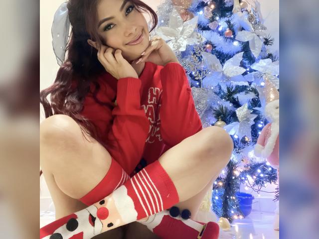 KendraMiler with long red hair, sitting, wearing red sweater, red socks with Santa face, showing her ass, hands on cheeks, smiling, Christmas tree with blue lights behind.