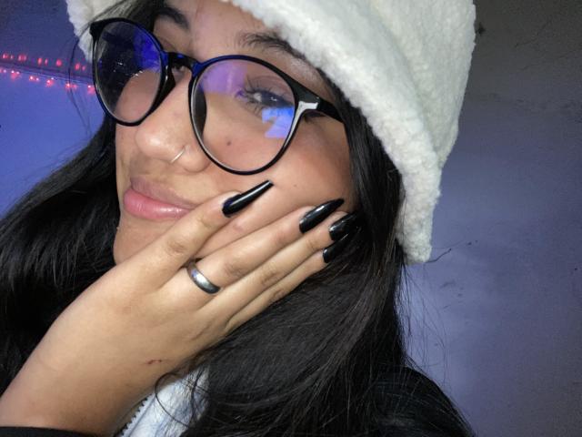 Elizaa, con le unghie nere, il berretto bianco e gli occhiali, che si tocca la guancia con un anello al dito, mostrando le sue labbra rosa e piene e il suo piccolo naso..