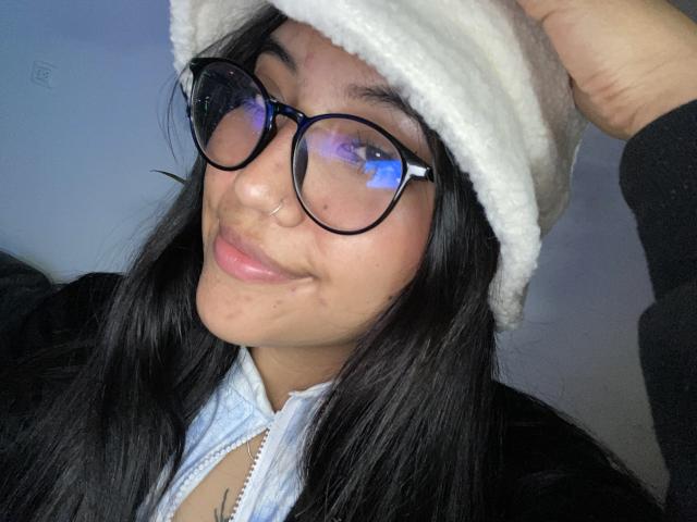 Elizaa, con occhiali rotondi, capelli neri, un cappello bianco e peloso, un anello al naso e una giacca azzurra con zip, sorride leggermente e tiene il cappello in alto. Le sue natiche non sono visibili..