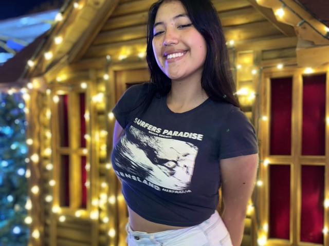 Elizaa, avec de longs cheveux noirs, souriante, portant un t-shirt gris moulant avec un motif blanc, et un pantalon blanc, se tient devant une maison en bois décorée de guirlandes lumineuses..