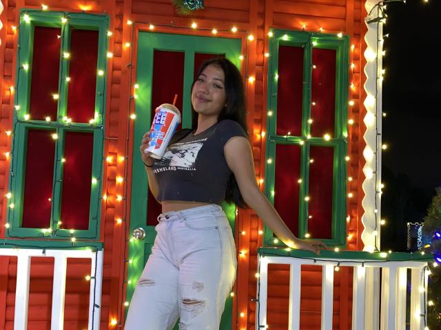 Elizaa with long black hair, holding a soda can, wearing a tight gray crop top and ripped white jeans, standing against a colorful, string-lit wooden structure, smiling.