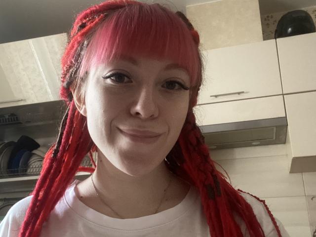 PassionSupply with red dreadlocks, white shirt, and slight smile, showing nose ring, eyeliner, and pink bangs, in a kitchen. She has fair skin and small nose.