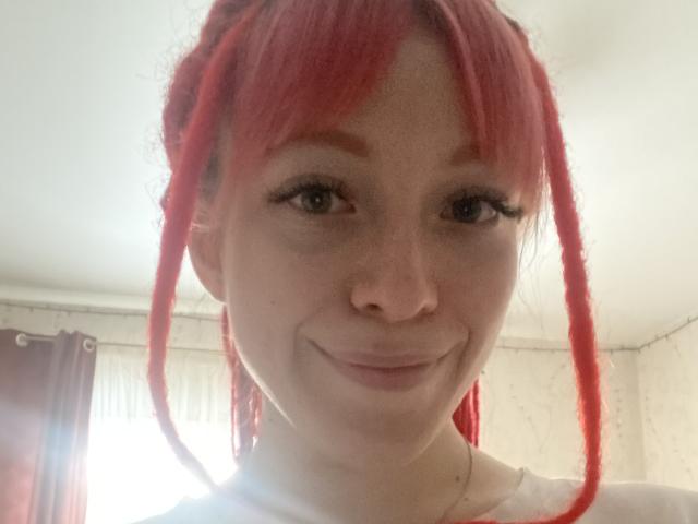 A PassionSupply tem dreadlocks vermelhas, um sorriso adorável e sardas no rosto. Ela está vestindo uma camisa branca, tem cílios longos e uma fonte de luz suave atrás dela..
