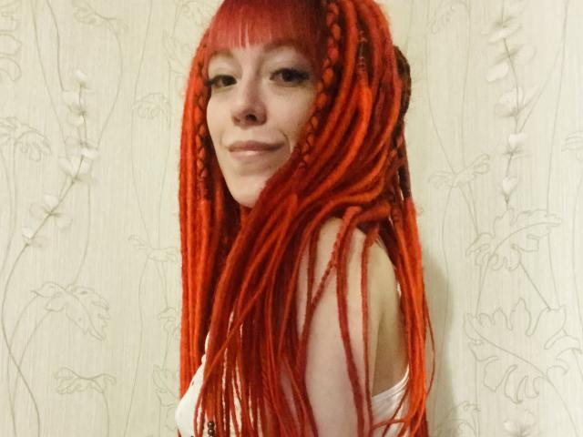 PassionSupply, com dreadlocks vermelhas vibrantes e longas, sorrindo para a câmera, vestindo uma camiseta branca, mostrando seus ombros nus e um pequeno piercing no nariz..