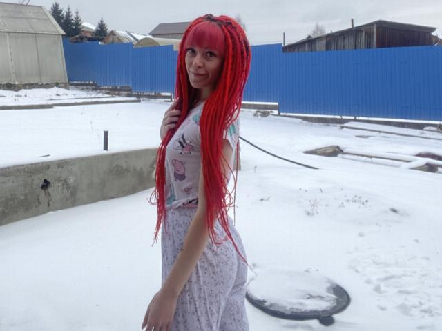 PassionSupply with bright red braided hair, small tits, and a cute smile, wearing a white floral dress, stands in a snowy yard with a blue fence.