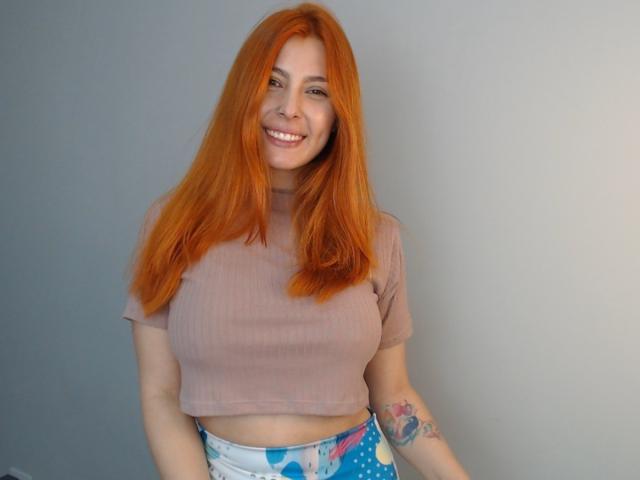 AnnyCraffort with red hair, smiling, wearing a pink ribbed crop top, blue floral pants, showing a tattoo on her right arm, standing against a plain gray wall.