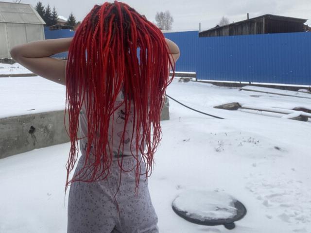PassionSupply with red braids, arms raised, wearing a white tank top and gray shorts, standing in a snowy yard with a blue fence and a round metal cover.