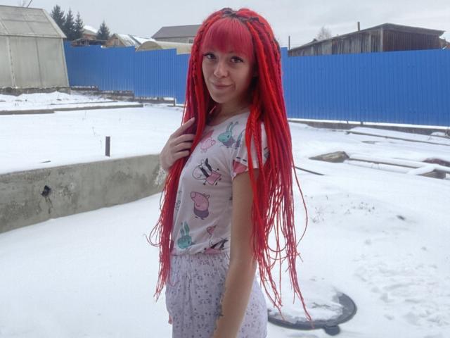 PassionSupply met felrode dreadlocks, kleine borsten en een schattige glimlach, gekleed in een roze shirt met een vlinderprint en lila broek, staand in een besneeuwde tuin met een blauwe omheining..