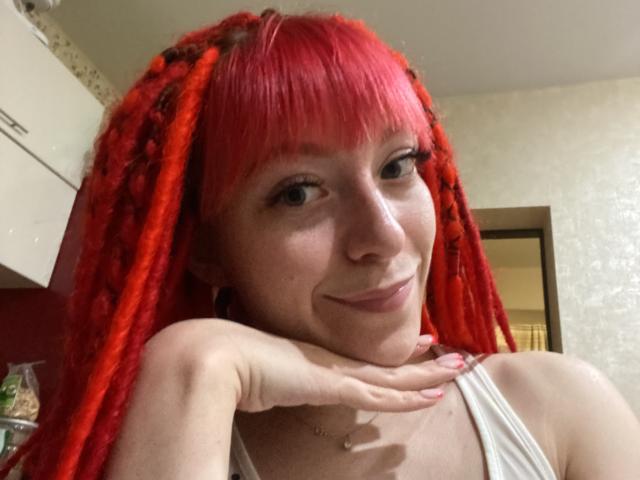 PassionSupply, com dreadlocks vermelhas, franja e um sorriso adorável, apoiando o queixo na mão, vestindo uma camiseta branca, mostrando um colar pequeno, em um selfie casual..