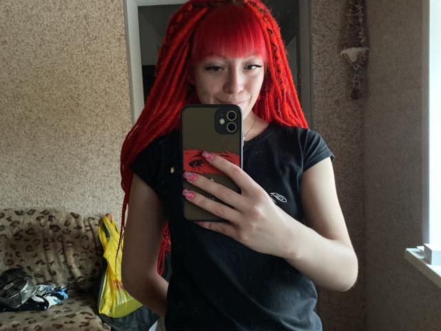 Uma selfie da PassionSupply com dreadlocks vermelhas vibrantes, vestindo uma camiseta preta, segurando um telefone, sorrindo, mostrando unhas cor-de-rosa, em um quarto com uma janela..
