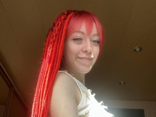 Mulher ruiva, PassionSupply, com marias-chiquinhas trançadas, nariz pequeno, sorrindo, vestindo uma camiseta branca sem mangas, luz do sol no rosto, close do tronco superior, teto com luz ao fundo..
