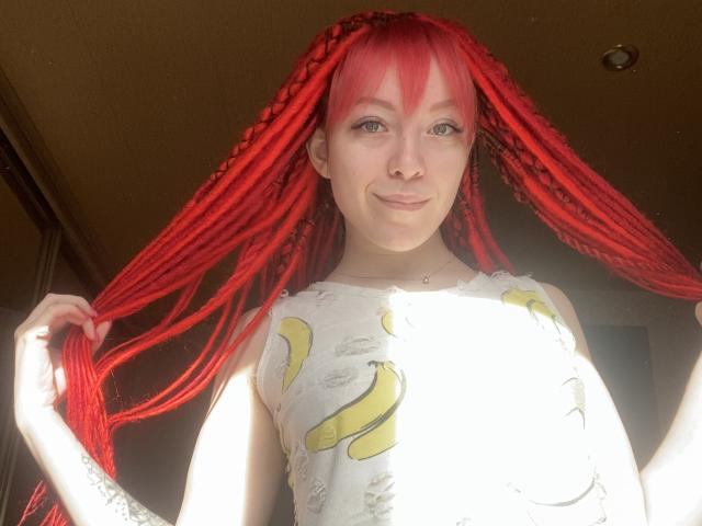 PassionSupply com dreadlocks vermelhas vibrantes, segurando-as, vestindo uma camisa com estampa de bananas, sorrindo, iluminada pelo sol, pequena perfuração no nariz, pele clara, olhos verdes, maquiagem discreta, luz vindo de cima..