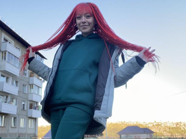 PassionSupply met felrood haar, een groene hoodie en groene broek, haar haar naar voren houdend, lachend, staand voor een gebouw, een grijze jas aan..