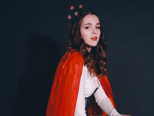 JuliaKing tem cabelos castanhos ondulados, batom vermelho e usa um capuz vermelho com pequenos enfeites de flores na cabeça. Ela está vestindo uma blusa branca e olhando para o lado..