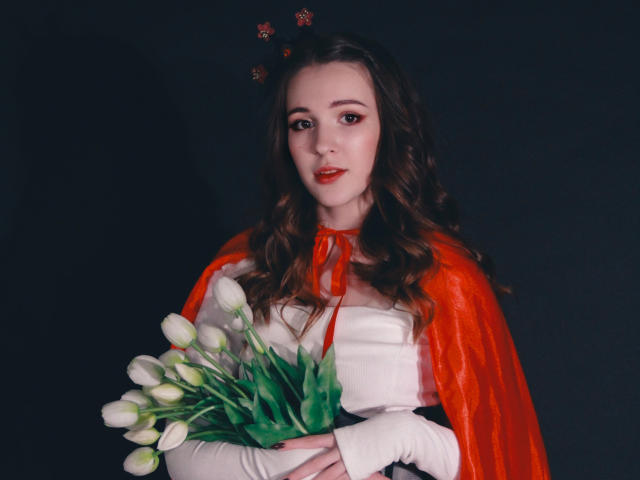 JuliaKing, avec de longs cheveux bruns ondulés, une cape rouge-orange, tenant des tulipes blanches, portant un haut blanc, sur un fond sombre, avec du rouge à lèvres, un léger sourire, un serre-tête floral rouge, et un éclairage doux..