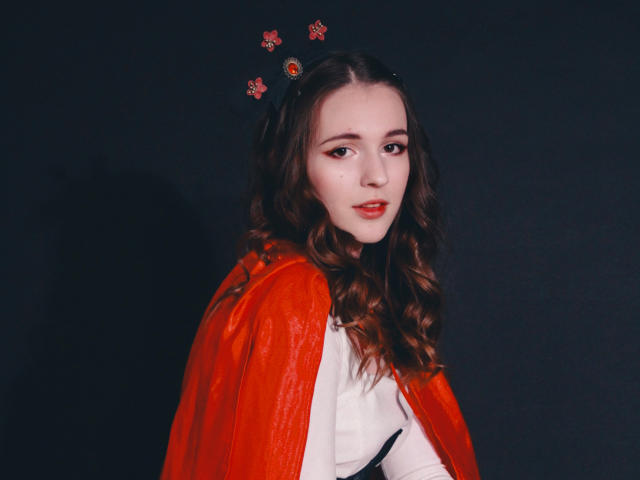 JuliaKing, avec de longs cheveux bruns ondulés, des yeux rouges et du rouge à lèvres rouge, portant une cape rouge transparente sur une chemise blanche, et un ornement de tête avec des motifs floraux rouges..
