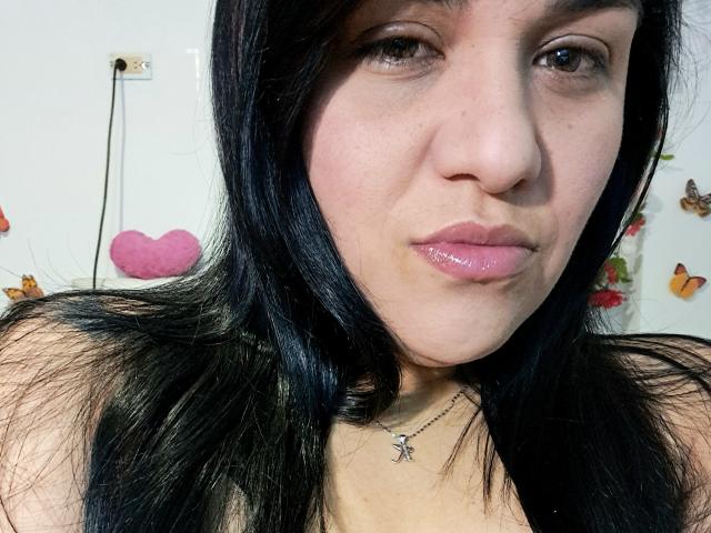 VivianaThomson has shiny pink lips, black hair, and a silver necklace with a butterfly charm. Her bare shoulders are visible. She's making a slight pouty face.