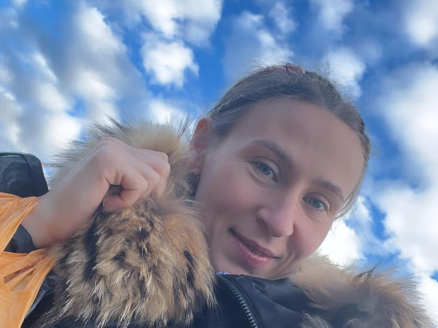 Saveta, met een schattende glimlach, draagt een behaarde jas en houdt de kraag vast met haar rechterhand. Op de achtergrond is een blauwe lucht te zien met pluizige wolken. Ze heeft bruin haar en een lichte huidskleur..
