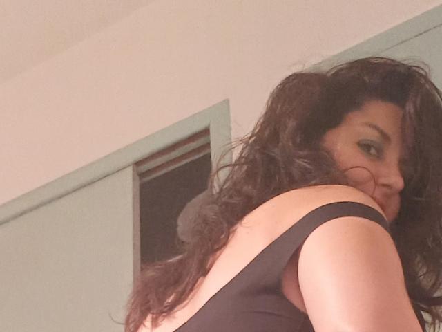 LaFrancaiseJessy, met krullend zwart haar, draagt een zwarte tanktop, waarbij haar schouder en rug zichtbaar zijn. Ze kijkt over haar schouder met een lichte glimlach..