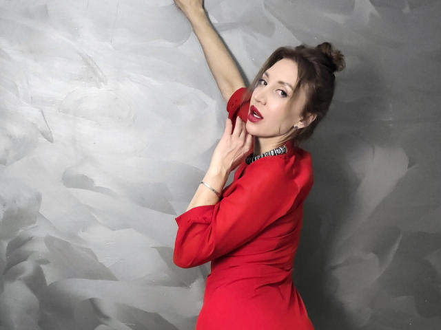 SimoniGrace com vestido vermelho, braços levantados, mão na parede, cabelo castanho preso em um coque, batom vermelho, olhando por cima do ombro, mostrando os seios, fundo de parede cinza com textura, boca ligeiramente aberta..