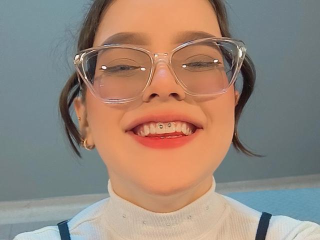 Kim, zomerse outfit met een bril met heldere glazen, lachend, met metalen beugels op haar tanden, draagt een witte coltrui, bruin haar in een staart, gouden oorringen en een blauwe band..