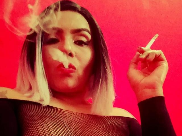 AbbyCrown con cabello de rubio a negro, fumando, sosteniendo un cigarrillo, vistiendo una blusa de malla negra, fondo rojo, expresión de labios fruncidos, humo visible, nariz ligeramente levantada, postura relajada..