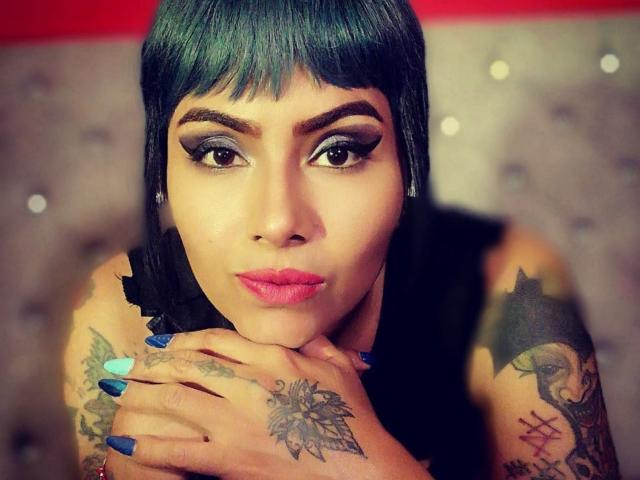 AbbyCrown mit einem türkisfarbenen Bob, starkem Eyeliner, pinkem Lippenstift, blauen Nägeln, Tattoos an Händen und Armen, einem schwarzen Top ohne Ärmel, die die Lippen zusammenpresst, sich auf die Hände stützt und in die Kamera schaut..